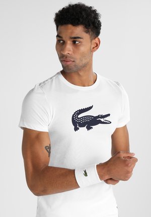T-shirt bianca in cotone con una grande grafica di un coccodrillo blu navy. Il design include motivi triangulari. È visibile un braccialetto con un logo verde.