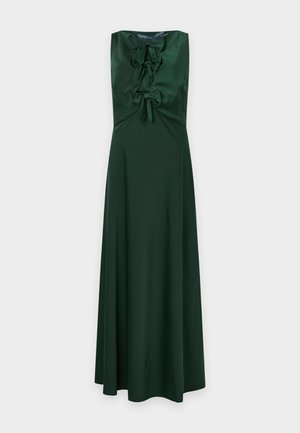 Une femme porte une robe sans manches vert foncé, à détails floraux et de longueur cheville. Elle tient une pochette marron et porte des chaussures à talons à brides.