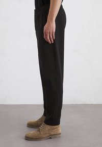 Black elastic-waist pants with side pockets, tapered shape, and smooth texture, paired with tan suede shoes featuring round toe and laces.