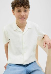Biała krótka koszula na guziki z teksturowanej tkaniny, w połączeniu z jasnoniebieskimi dżinsami. Model siedzi na drewnianym krześle.