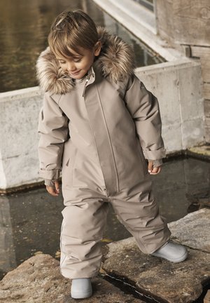 Beige geïsoleerde sneeuwpak met een met bont gevoerde capuchon, elastische mouwen en een ritssluiting aan de voorkant, gecombineerd met lichtgrijze waterdichte laarzen.