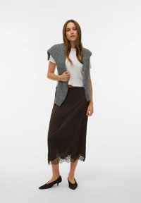 Chaleco gris de punto sobre una camiseta blanca corta, falda marrón de satén con ribete de encaje y zapatos negros de tacón bajo. Conjunto sencillo y en capas.