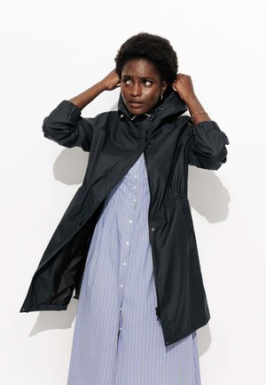 Veste noire imperméable avec capuche, taille cintrée et poignets élastiques, portée par-dessus une robe rayée bleue et blanche. Texture lisse.