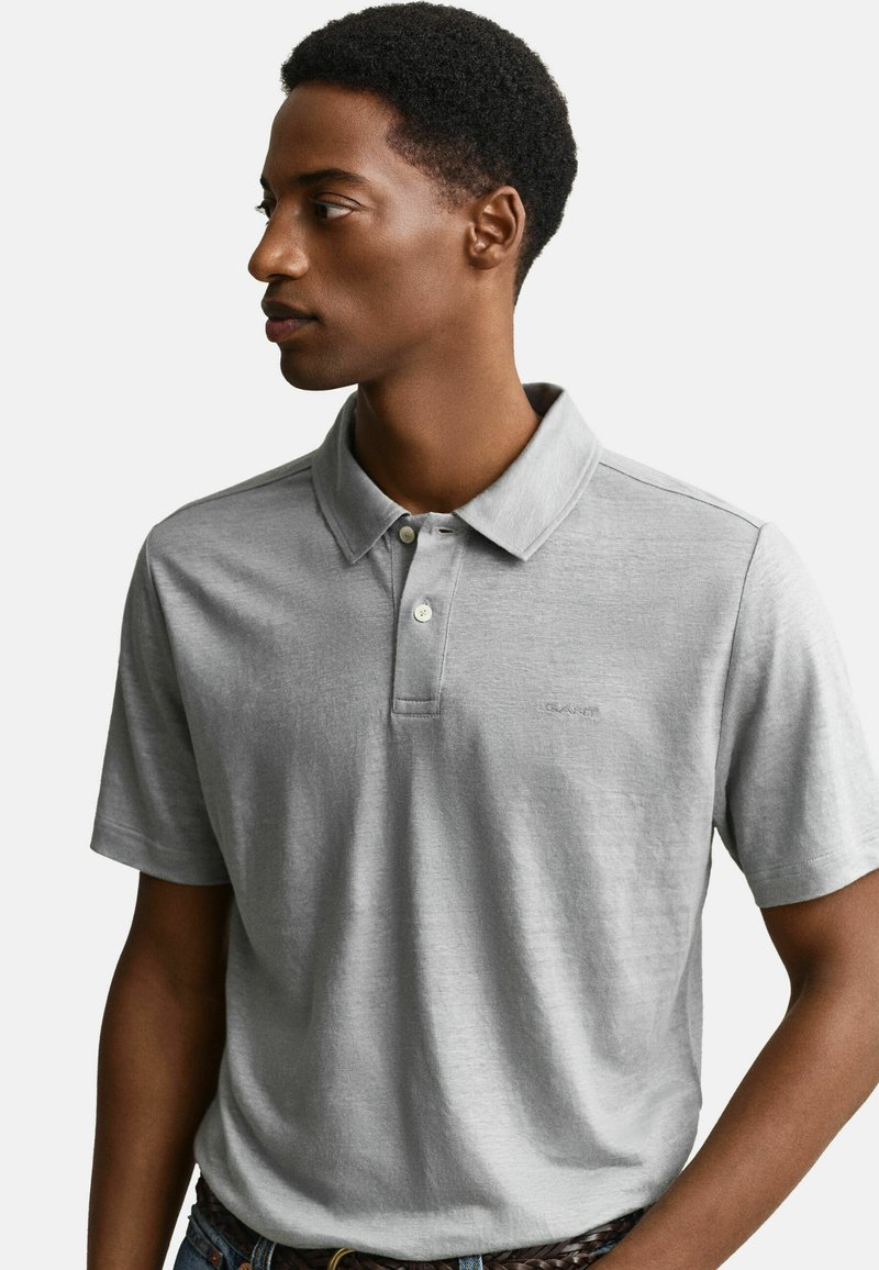 Junger Mann in einem hellgrauen kurzärmeligen Poloshirt mit zwei Knöpfen, der nach links schaut vor einem schlichten hellen Hintergrund.