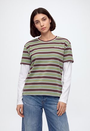 Le mannequin porte un T-shirt rayé à manches courtes par-dessus un haut blanc à manches longues avec un jean bleu ample sur un fond de studio uni.