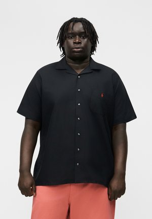 Άνδρας με μεσαίου μήκους dreadlocks, που φοράει μαύρο κοντομάνικο πουκάμισο με κουμπιά και κοραλλί σορτς, στέκεται μπροστά σε ένα απλό φόντο.