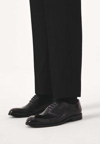 Čierne kožené spoločenské topánky so špicatou špičkou, viazaním na šnúrky, lesklým povrchom a zdvojeným opätkom, v kombinácii s priliehavými čiernymi nohavicami.