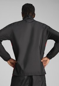 Veste légère noire avec un col haut, présentant une texture lisse et un détail d'accent rouge sur le côté, vue de dos.