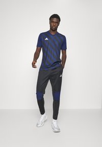 Marineblaues und schwarzes Sportoutfit bestehend aus einem gestreiften Kurzarmshirt und tapered Pants mit blauen Akzenten, kombiniert mit weißen Sportschuhen.