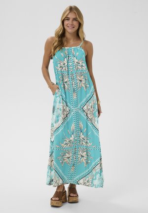 Nő türkiz színű, virágos mintájú, spagettipántos maxiruhában és barna platform szandálban, kezét a zsebében tartva, mosolyogva áll.