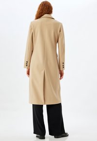 Cappotto in lana beige con taglio sartoriale, colletto a rever, spacco singolo sul retro e tre bottoni neri su ciascun polsino. Abbinato a pantaloni neri.