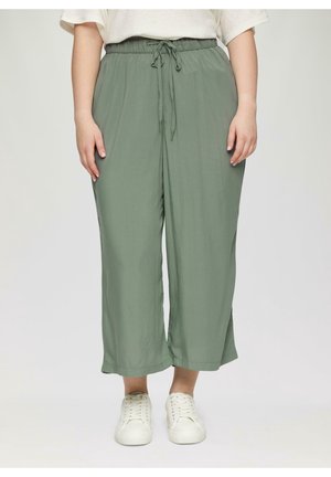 Persoon die losse, saliegroene broek met trekkoord en witte sneakers draagt, staand tegen een effen lichte achtergrond.