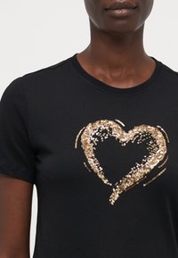 Černé tričko s krátkým rukávem, které má zlatý design srdce z flitrů. Měkká látka s kulatým výstřihem, která se vyznačuje hladkou texturou a lesklým akcentem.