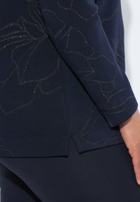 Maglietta a maniche lunghe navy con motivo floreale testurizzato in oro metallico. L'orlo presenta una fessura laterale, aggiungendo dimensione al design.
