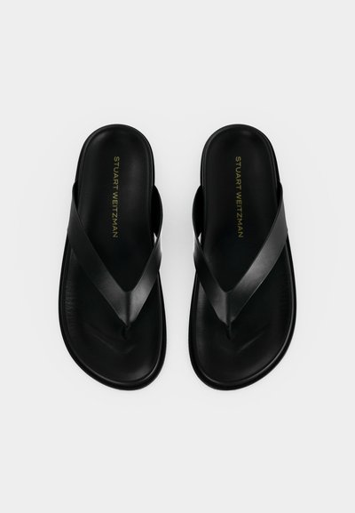 Μαύρα δερμάτινα σανδάλια flip-flop με φαρδιές τιράντες και επίπεδες σόλες, με το λογότυπο "Stuart Weitzman" τυπωμένο με χρυσό στα πατούμενα, εμφανιζόμενα από πάνω.