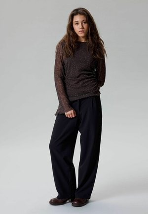 Fiatal nő hosszú hajjal, fekete áttetsző hosszú ujjú felsőt, bő fekete nadrágot és barna cipőt viselve, egy egyszerű háttér előtt áll.