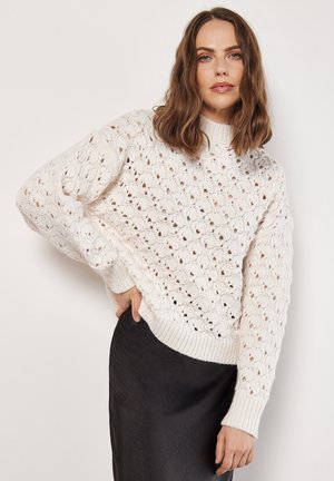 Weißer, locker sitzender Strickpullover mit einem strukturierten, durchbrochenen Muster und gerippten Bündchen. Kombiniert mit einem schwarzen Rock.