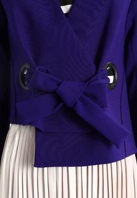 Fialová bunda s překládaným designem, vybavená předním zavazováním na mašličku, velkými černými průchodkami a plisovanou bílou spodničkou. Hladká textura.