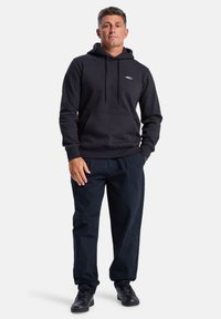 Black hoodie with front pouch pocket and drawstrings, paired with dark pants and black shoes. Features a small logo on the chest.