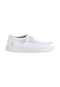 Zapato tipo slip-on de lona blanca con parte superior texturizada, detalles de cordones y suela de goma de color claro. Diseño minimalista sin patrones visibles.