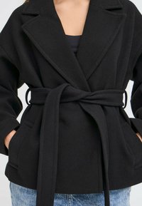 Donna che indossa un cappotto di lana nero con cintura, ampi risvolti e tasche, abbinato a jeans di denim blu.