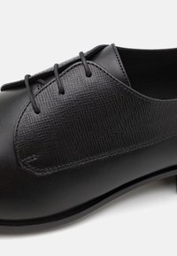 Black leather shoe featuring a textured panel, rounded toe, and four eyelets for laces, with a smooth finish and sturdy sole.