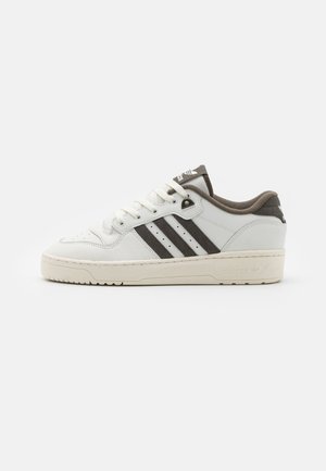 Weißer flacher Adidas Sneaker mit drei schwarzen Streifen, Schnürung vorne und beiger Sohle auf einem einfarbigen hellen Hintergrund.