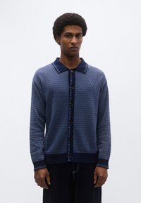 Καζάκα σε ναυτικό μπλε με γιακά, υφή υφάσματος, κλείσιμο με κουμπιά και αντίθετο ριγέ σχέδιο στα τελειώματα και στην περαστή τιράντα.