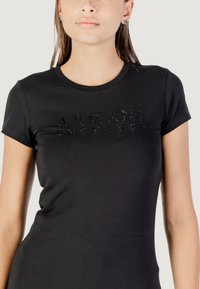 Camiseta negra de manga corta con cuello redondo. Presenta un logo texturizado de "ARMANI EXCHANGE" en brillantes tachuelas en el pecho. Tejido suave.