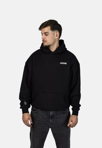 Černá oversized mikina s kapucí se šňůrkami, přední kapsou a bílým logem. Kombinována s tmavými džínami a jemnými detaily na manžetách.