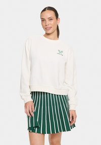 Sweatshirt crème à manches longues et ourlet côtelé, présentant un graphique de raquette de tennis verte et le texte "racket club", associé à une jupe plissée rayée verte.
