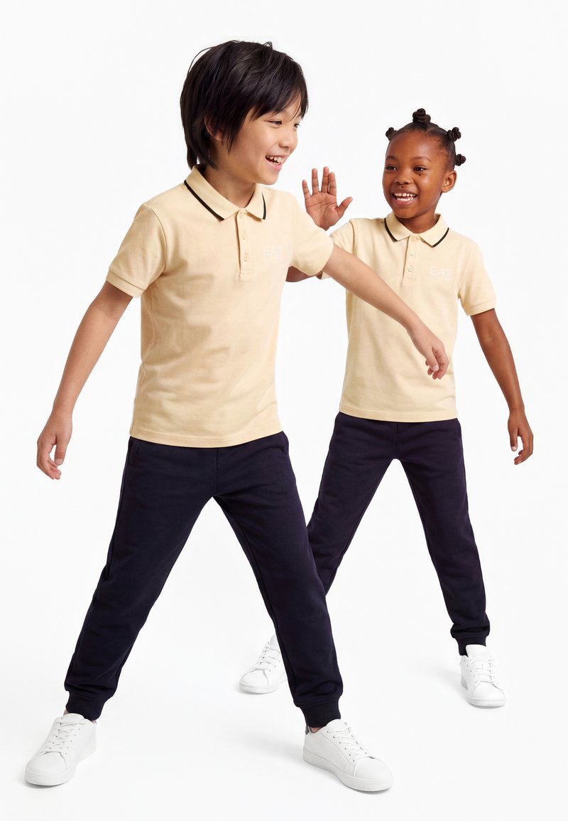 Deux enfants en polos beige et pantalons foncés sourient et jouent, les bras tendus, sur un fond blanc.