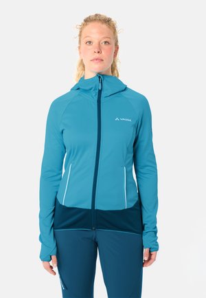 Femme blonde portant une veste zippée bleue Vaude avec capuche et un pantalon bleu assorti, debout devant un fond blanc uni.