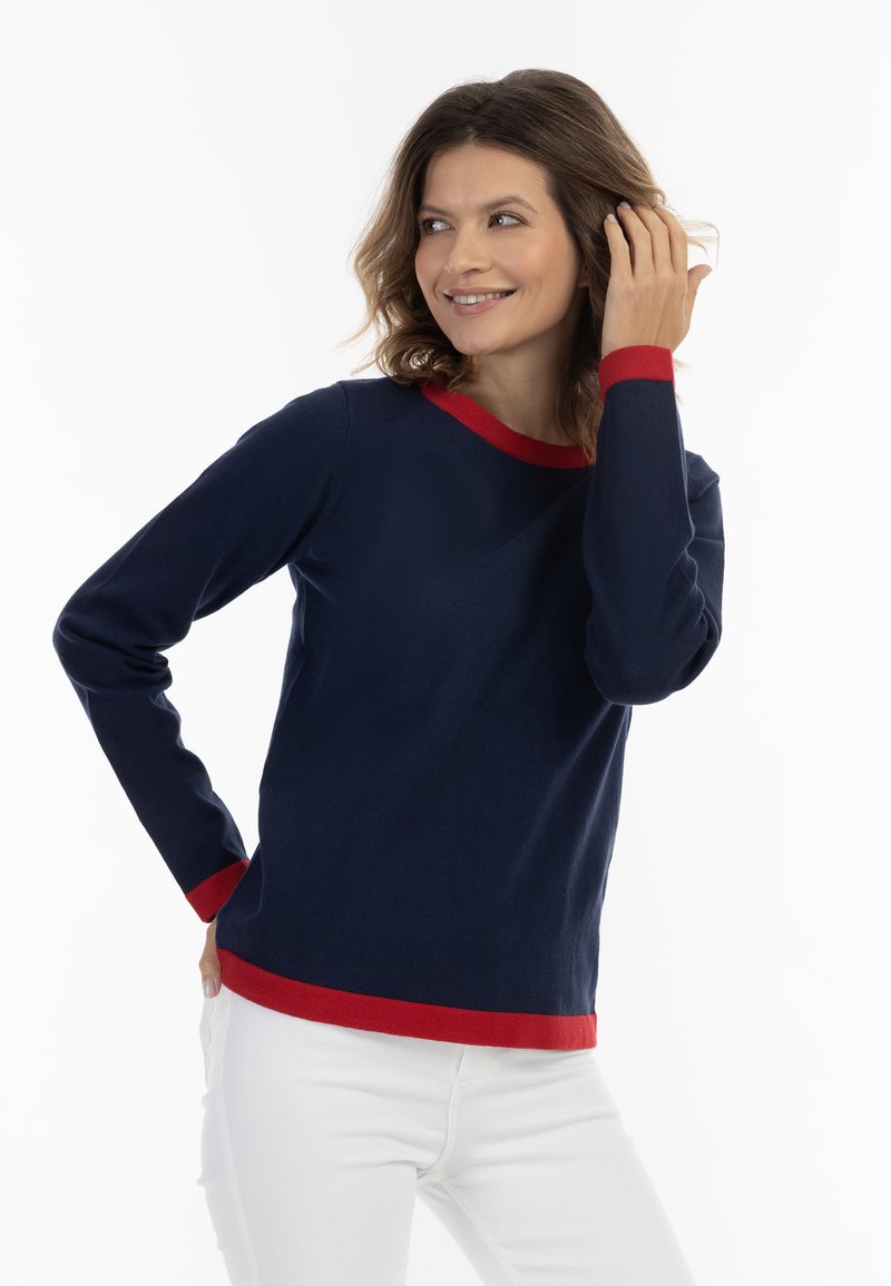 Pulover marin cu guler și manșete roșii, mâneci lungi, textură moale, gât rotund, asortat cu pantaloni albi. Fără modele sau elemente metalice vizibile.
