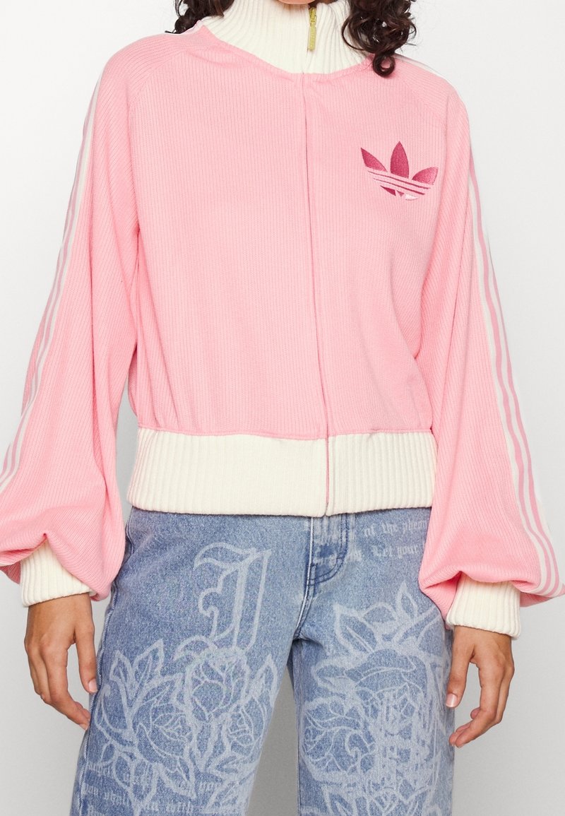 Rosa geripptes Reißverschluss-Jacke mit cremefarbenen Akzenten, ausgestattet mit drei weißen Streifen an den Ärmeln und einem kleinen Logo auf der Brust.