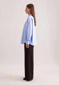 Hellblaues oversized Hemd mit hohem Kragen und langen Ärmeln, kombiniert mit maßgeschneiderten schwarzen weiten Hosen. Glatte Stofftextur.