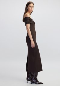 Braunes geripptes Off-Shoulder-Kleid mit seitlichem Schlitz und dekorativem Detail am Ausschnitt, getragen mit knielangen, glänzenden braunen Stiefeln.