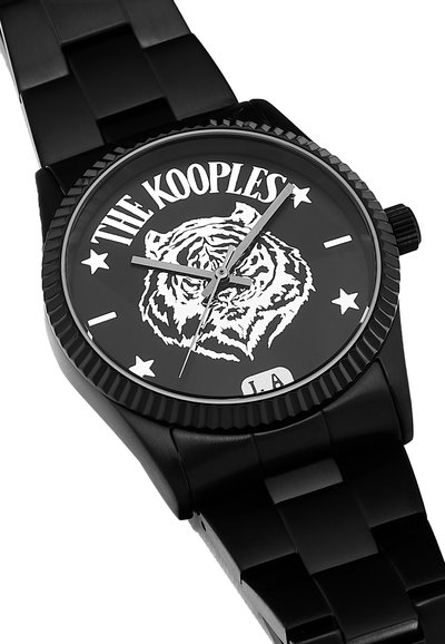 Reloj analógico negro con un bisel texturizado, que presenta un gráfico de tigre, estrellas y el texto "THE KOOPLES" en una esfera negra, con una correa de metal negro.