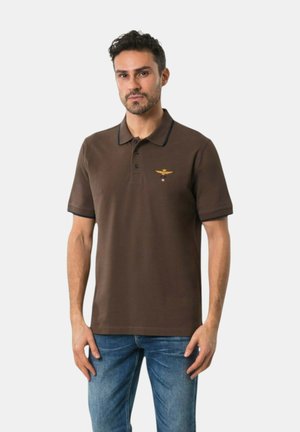 Uomo che indossa una polo marrone a maniche corte con bordi neri e uno stemma giallo ricamato sul petto, abbinata a jeans blu.