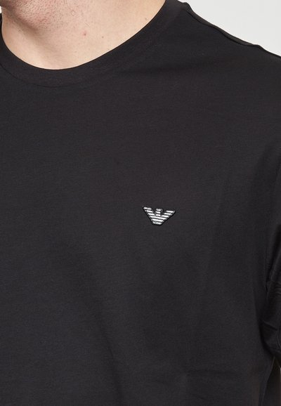 T-shirt en coton noir avec un col rond, arborant un logo brodé argenté sur la partie gauche de la poitrine. Texture lisse et design ajusté.