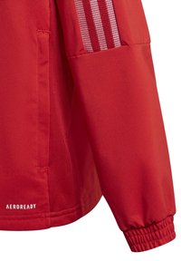 Rød sportsjakke med en glat tekstur, der har mesh-detaljer og tre lodrette striber. Elastiske manchetter og "AEROREADY" branding nederst.