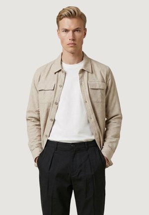 Ung mand med blondt hår iført en beige skjorte med knapper over en hvid t-shirt og sorte plisserede bukser, hænderne i lommerne, neutral baggrund.