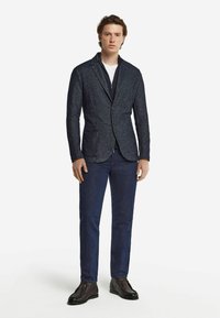 Marinblå texturerad kavaj med två framfickor, enknappsstängning och sjalkrage, tillsammans med mörka jeans och bruna skor.