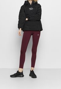 Personne portant une doudoune noire, des leggings Reebok bordeaux et des baskets noires, debout devant un fond gris uni.