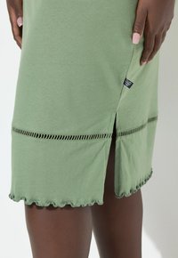 Vihreä puuvillamekko, jossa on halkio helmassa, jossa on aaltoileva reuna ja koristeelliset aukot. Sivusaumassa näkyvä etiketti.