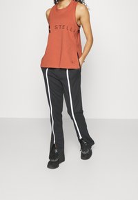 Rostfärgad ärmlös tanktop med svart logotryck, ihop med svarta byxor med vita dragkedjor på sidorna och en texturerad yta.