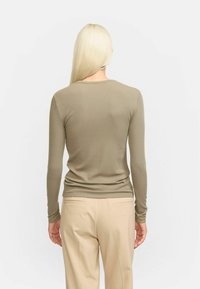 Cămașă lungă de culoare verde măslinie, cu guler rotund și siluetă ajustată, asortată cu pantaloni bej, croiți, cu buzunare pe spate.