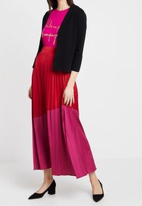 Kobieta ubrana w czarny krótki kardigan, różową koszulkę z grafiką, plisowaną spódnicę w czerwono-różowe pasy oraz czarne niskie obcasy, stojąca na tle białym.