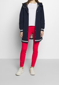 Marinblå vattentät parka med huva, silverdetaljer och tryckknappstängning, ihop med ljusröda slim-fit byxor och lätta sneakers.