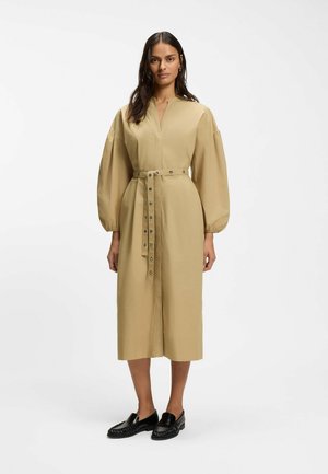 Kvinde stående iført en beige midi-kjole med lange ærmer og bælte, kombineret med sorte loafers mod en ensartet hvid baggrund.